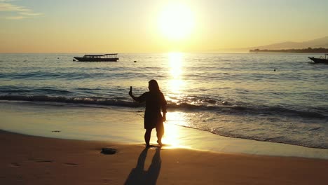 Girl-taking-selfie-photos-with-smartphone-on-empty-beach-at-sunset-golden-hour-with-orange-sky-reflecting-on-sea-surface