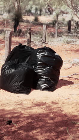 three black garbage bags sit on red dirt in a rural setting.