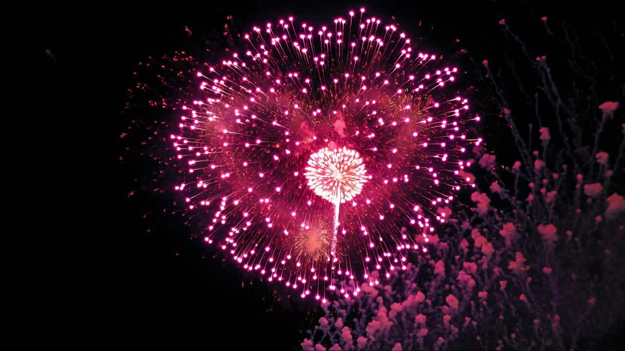 Aerial video captures vibrant pink fireworks exploding against a dark sky, with a low-angle shot