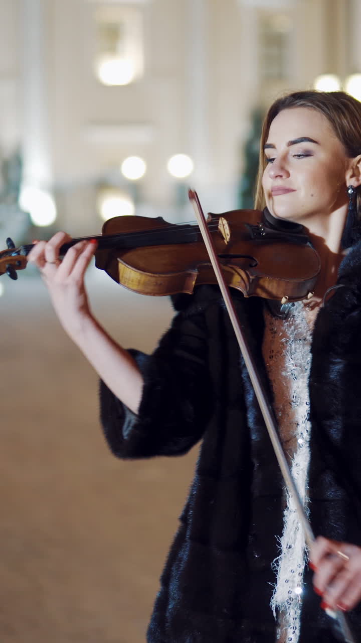 A young girl in a black coat is playing the violin on the background of buildings and evening lights in the centre of the city in the winter. Blurred Background Vertical video