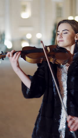 a young girl in a black coat is playing the violin on the background of buildings and evening lights in the centre of the city in the winter. blurred background vertical video