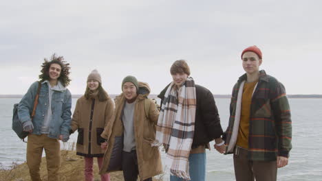 group of teenage friends in winter clothes are holding hands and raising arms on top of the mountain on the beach on a windy day