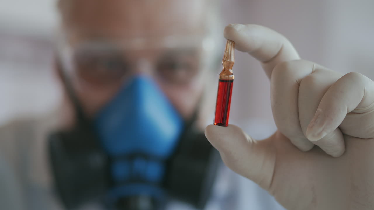 primer plano extremo de un científico con un respirador azul y gafas de protección un desarrollador de una vacuna contra el coronavirus con una ampolla roja. el médico mira la nueva vacuna. un nuevo tipo de medicamento contra el virus. imágenes de alta calidad 4k