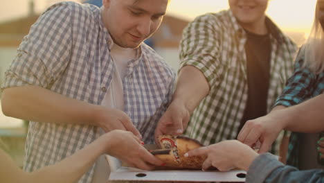 A-company-of-six-young-people-parses-pieces-of-hot-pizza-together.-This-is-a-rooftop-party-with-a-beer.