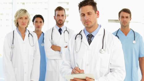 serious doctor writing on clipboard while staff are standing behind him