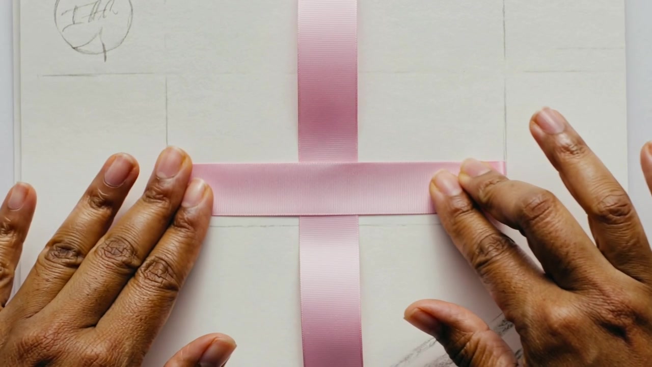 Placing adult hands folding and smoothing pink ribbon on white notebook, forming cross shape