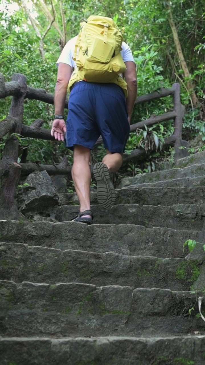 Tela vertical. Caminhante com mochila subindo degraus de pedra com mochila amarela, subida constante ao longo de um corrimão de madeira através de uma floresta verde densa perto de Da Nang, exploração ativa ao ar livre, sola visível.