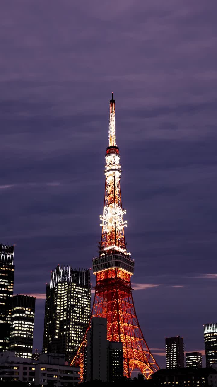 A low-angle shot of a brightly lit tower against a twilight sky, surrounded by city buildings