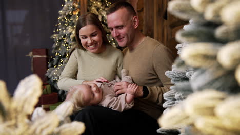smiling caucasian parents sit with their child. happy family of three portrait. christmas decorations at backdrop.