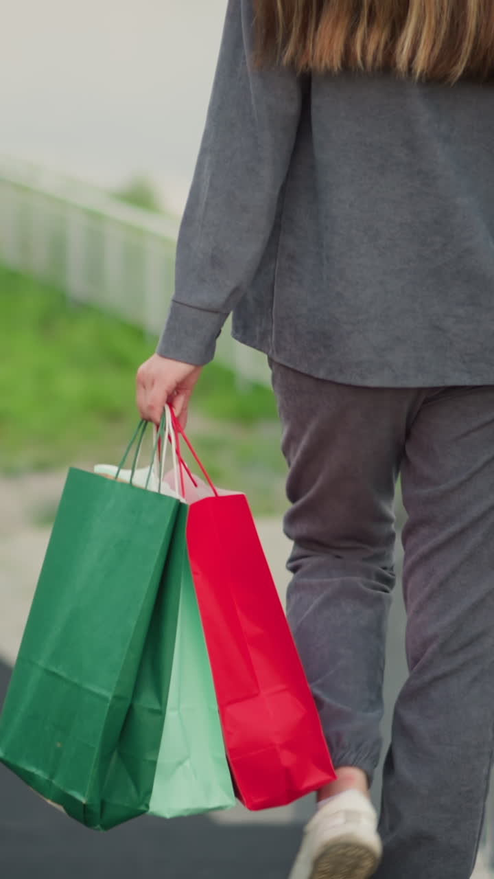 vista trasera de una mujer con ropa gris sosteniendo bolsas de compras coloridas mientras baja las escaleras, agarrando la barandilla con la mano para obtener apoyo, con vegetación borrosa en el fondo