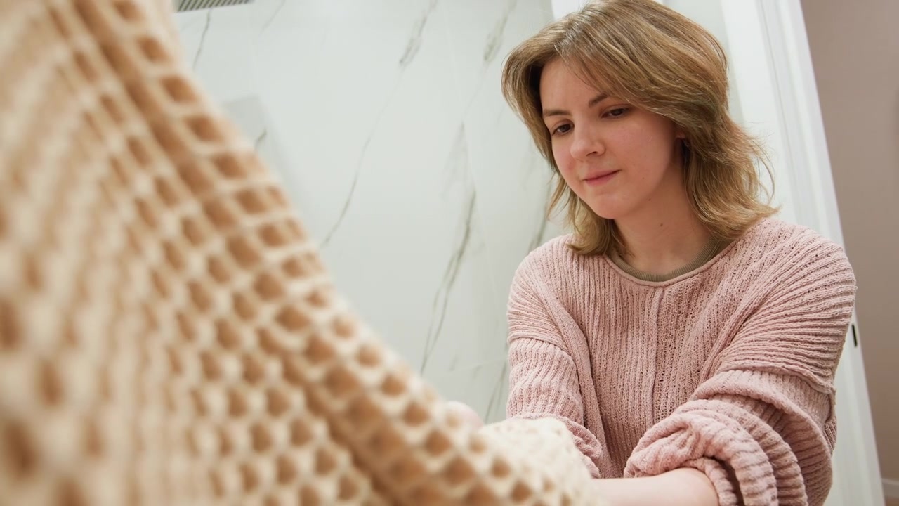 Young woman leans forward and reaches with cupped hands to help adjust clothing for another person in patterned robe, creating calm personal styling moment in bright marble dressing room with soft