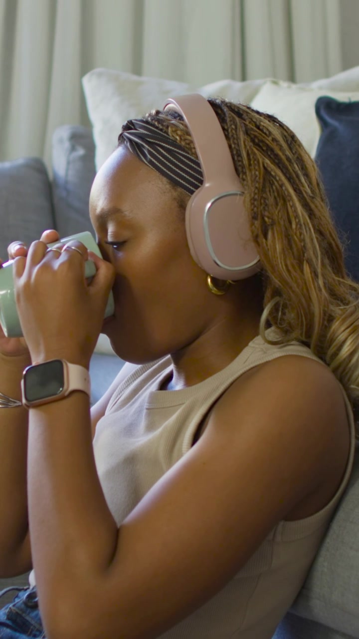 Vertical video: Finishing mug, woman in tan top grabbing remote on sofa, dozing with headphones