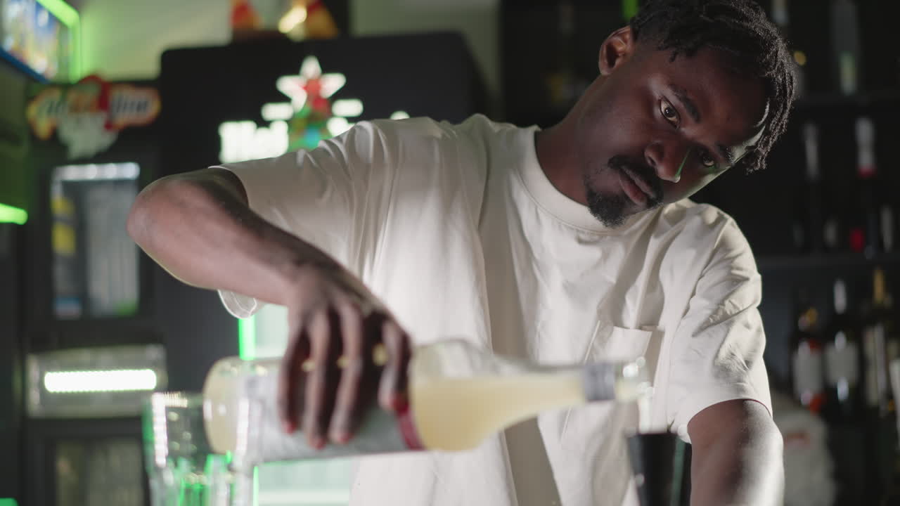 Bartender pouring liquid from bottle into glass at bar counter, focused on precise drink creation, with vibrant green neon lights in background enhancing bar ambiance