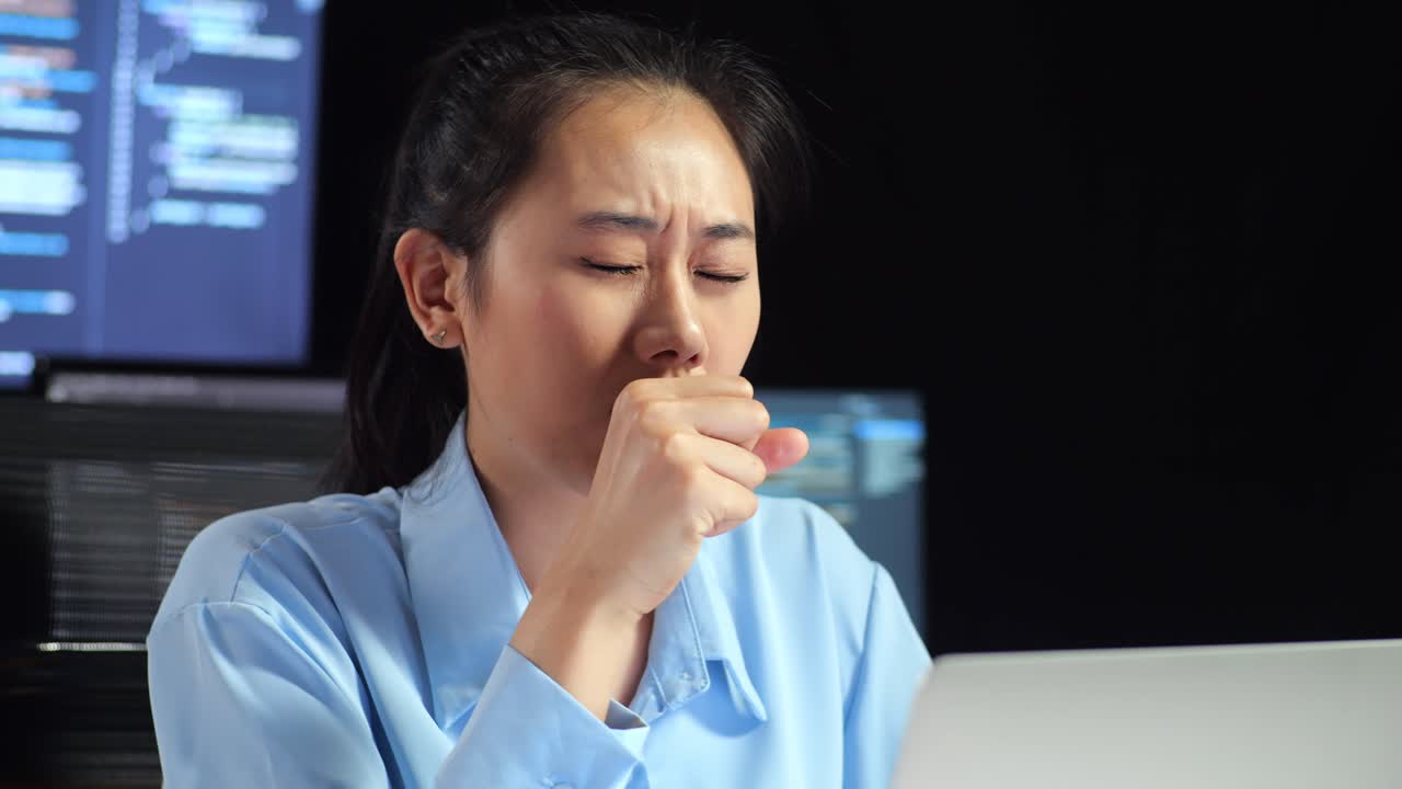 Close Up Of Asian Female Programmer Yawning While Writing Code By A Laptop Using Multiple Monitors Showing Database On Terminal Window Desktops In The Office