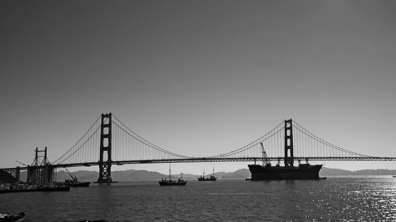 Black and white video still of a suspension bridge with ships, captured from a low angle