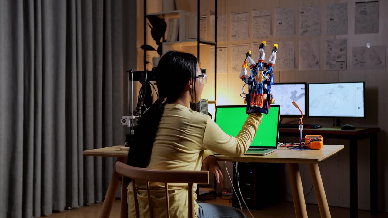 십대 아시아 소녀가 집에서 녹색 화면 모형을 가진 노트북에서 사이보그 손을 설계하는 것을 확인하는 뒷면