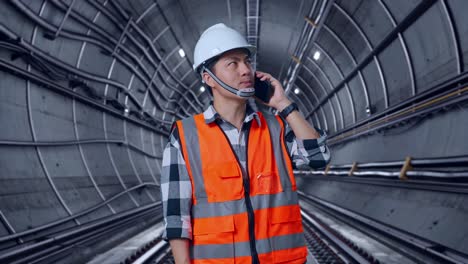asian male engineer with safety helmet talking on smartphone while standing in underground subway tunnel