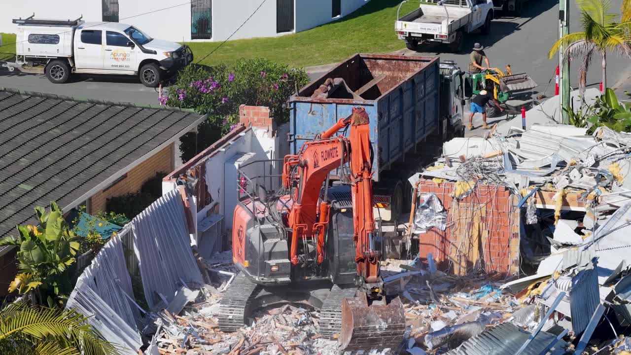 An orange excavator demolishes a residential house, clearing debris into a large dumpster under bright daylight at a suburban Gold Coast location. Static elevated camera view