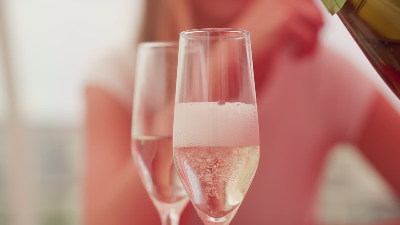 Due calici da champagne in primo piano con una bottiglia che versa spumante, donna sfocata con camicia rosa sullo sfondo, appuntamento intimo su un balcone al tramonto, spuma in aumento e bollicine scintillanti, bokeh delicato e luce calda