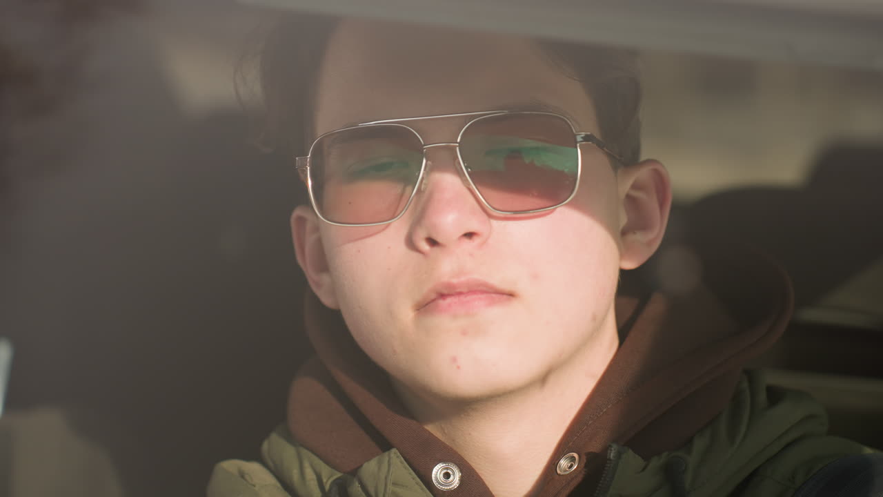 close up driver in winter jacket and hoodie wearing tinted glasses with sunlight reflections on face while leaning forward in car cabin, steady gaze through windshield, hand gripping steering wheel