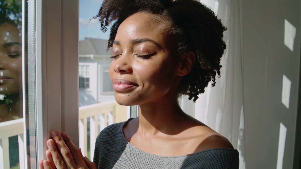 Standing woman pressing hands on balcony door, pausing as sunlight streaming on off-shoulder top