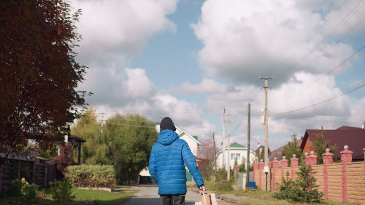 푸른 후디와 검은 모자를 입은 어린 소년이 오른손에 종이 가방을 흔들며 주거 거리를 고 있는 뒷면, 가을 잎자루와 구름이 가득한 하늘 아래의 다채로운 집들