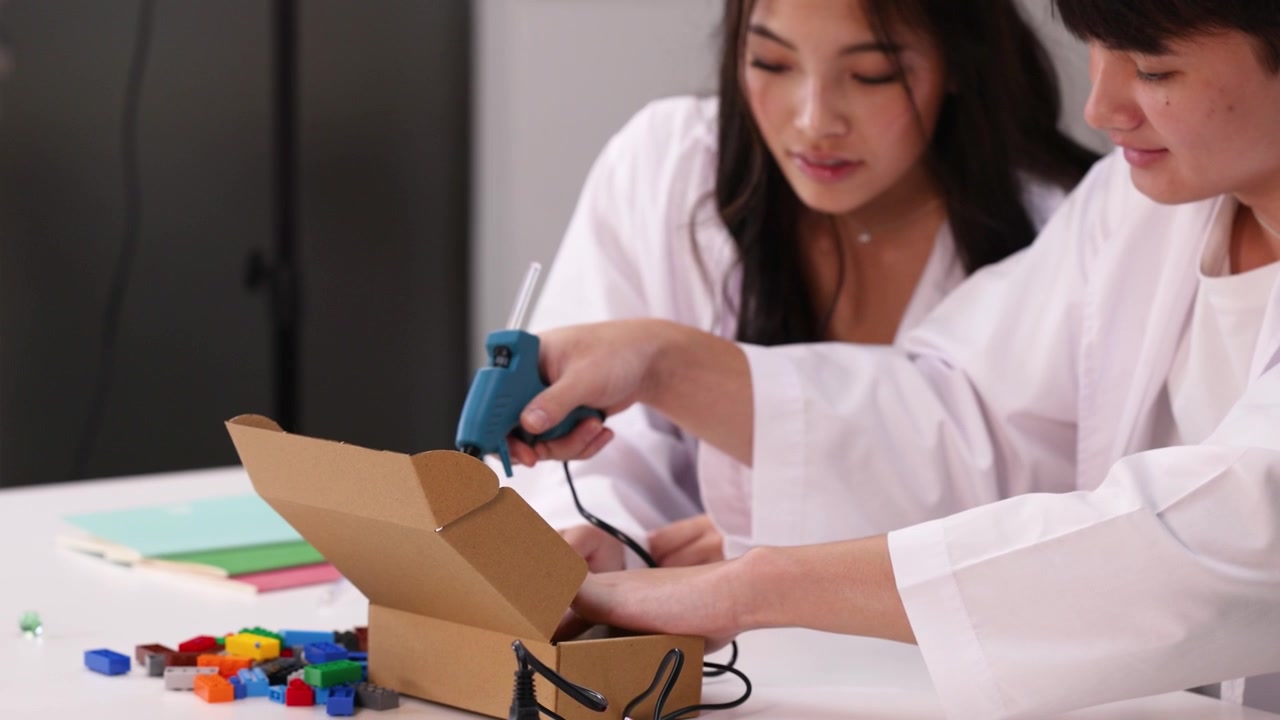 Asian teenagers in lab coats assemble a cardboard project using a hot glue gun.