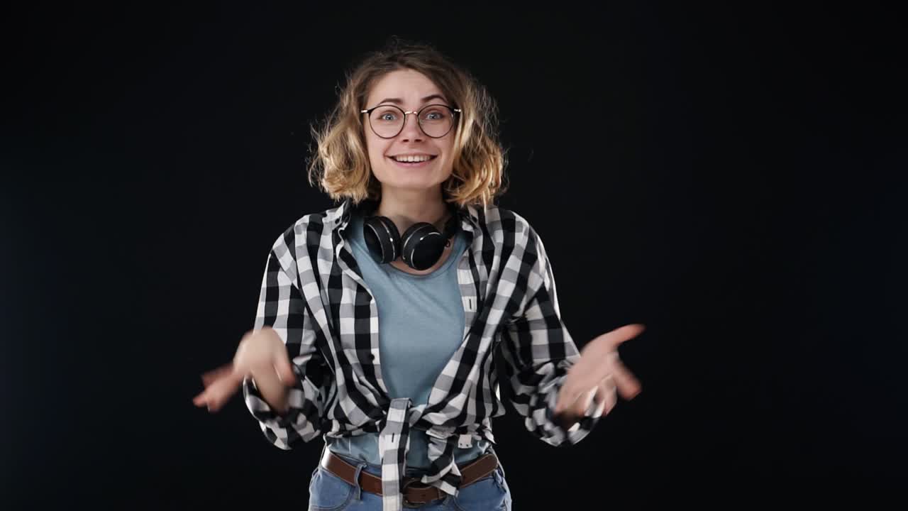 Over excited female with curly hairstyle, laughs happily, expresses sincere emotions, being surprised by good news. Happy young female in plaid shirt and jeans gesturing, super surprised over black background