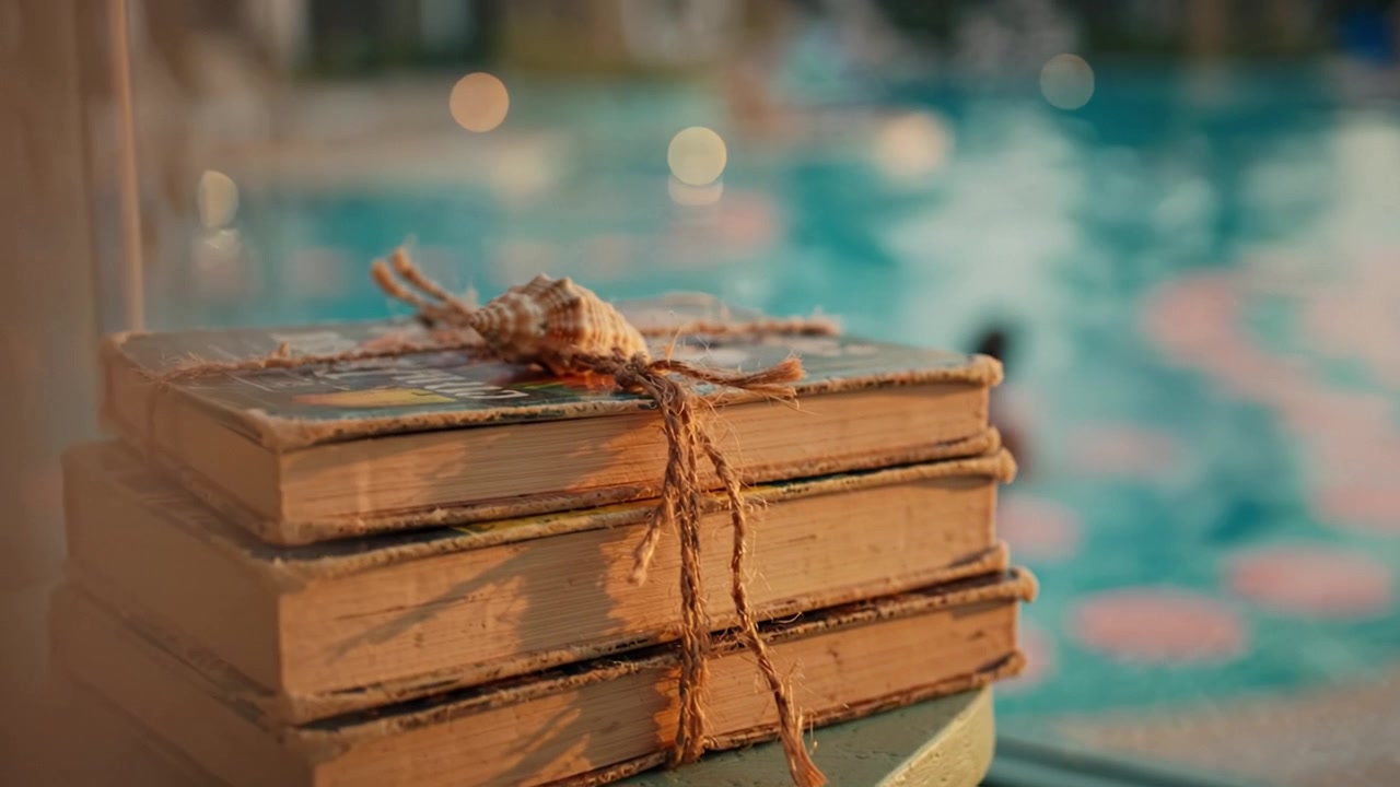 Scena serena a bordo piscina con libri vintage impilati e legati con spago, su uno sfondo sfocato di una piscina rilassante e lievi increspature dell'acqua.