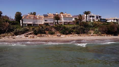 Vista-Aérea-De-Las-Olas-Del-Mar-Rompiendo-En-La-Playa-Con-Vistas-A-Las-Villas-De-Vacaciones-En-Estepona,-Costa-Del-Sol