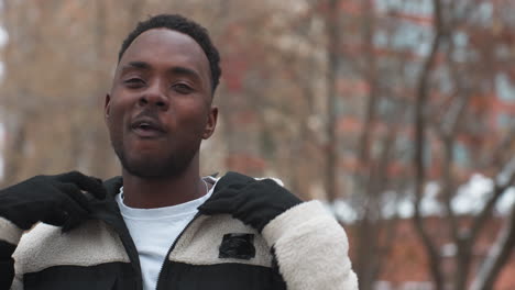 stylish black man removes hoodie while chewing gum, posing confidently in outdoor winter setting with bare trees in background, embodying casual fashion and urban lifestyle