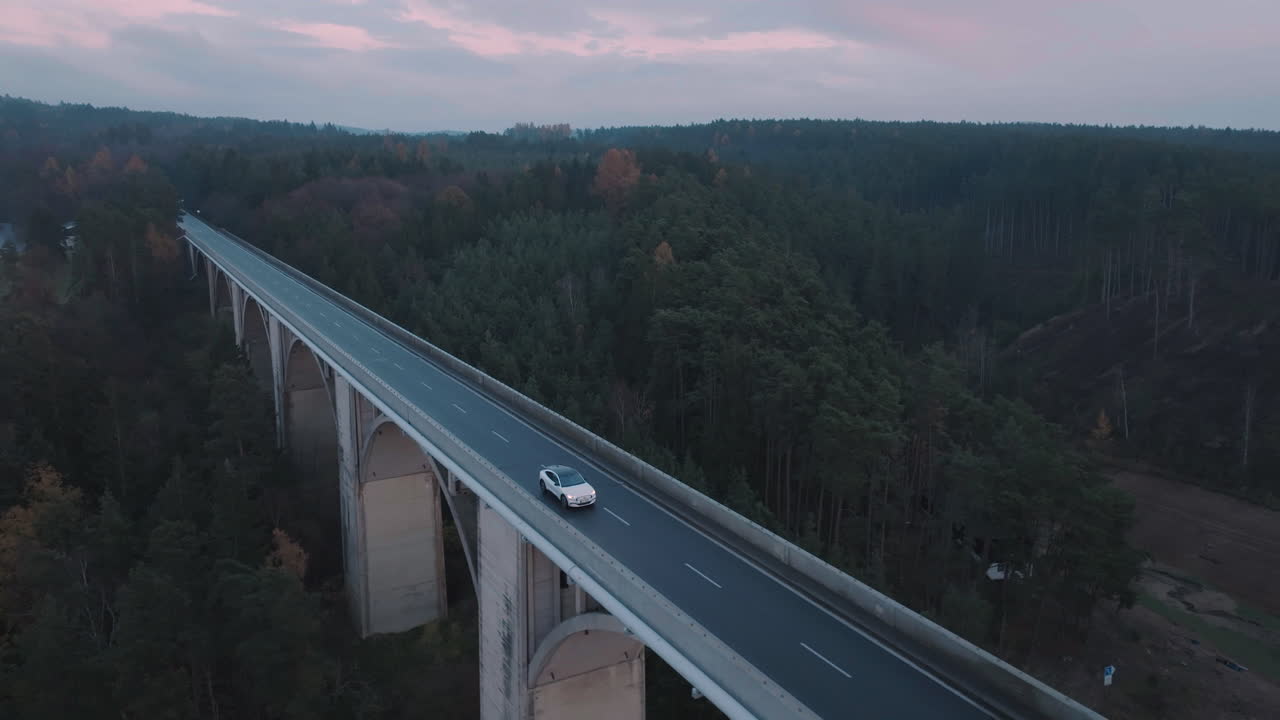 Veduta aerea di un'auto elettrica bianca in movimento su una strada di ponte vuota al crepuscolo, ripresa drone in tracking