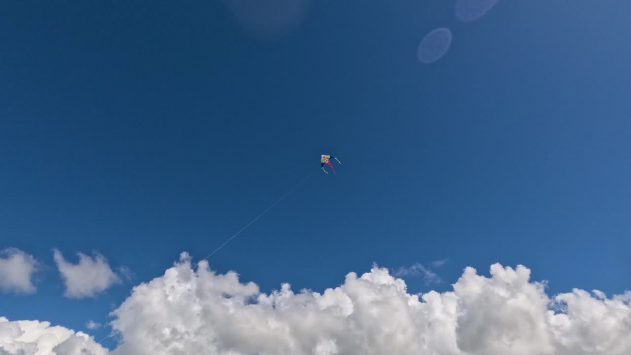 Un aquilone vola alto nel cielo sopra una spiaggia. Il sole splende luminoso mentre le nuvole si muovono lentamente sullo sfondo. La gente si gode l'aria aperta e osserva l'aquilone.