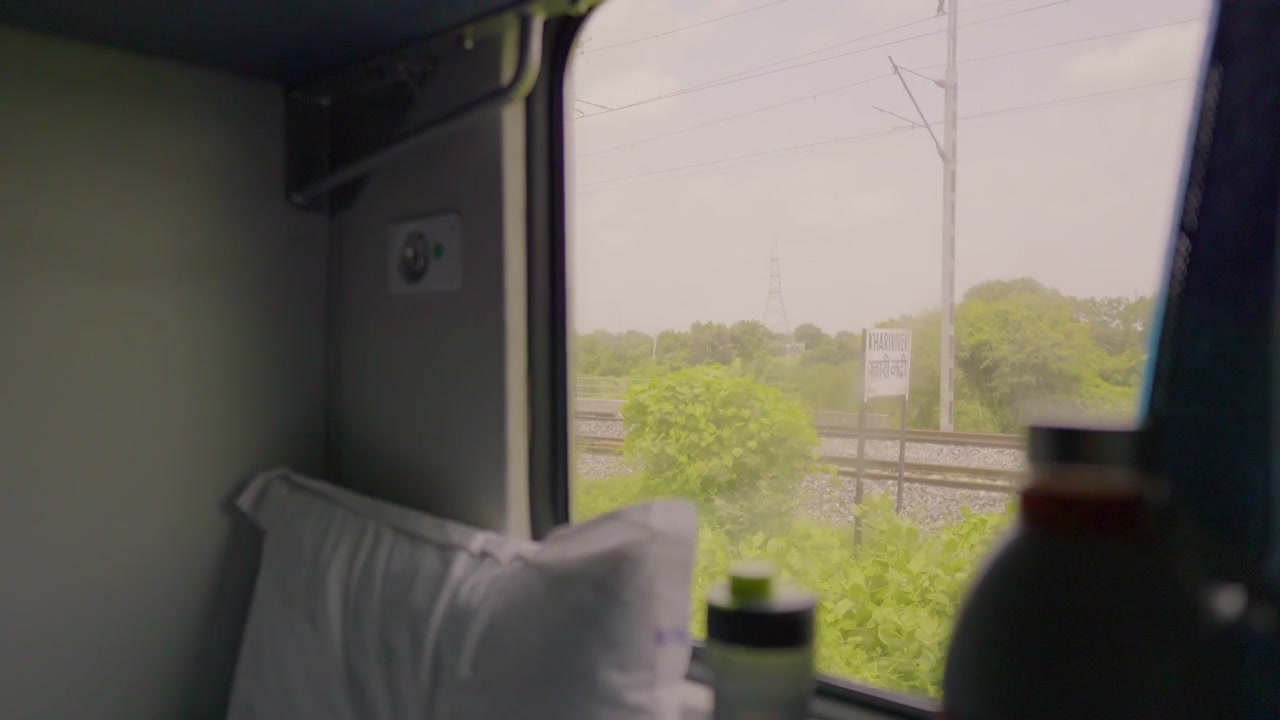 Guardando dal finestrino di una cabina di un treno passeggeri climatizzata la campagna verde che scorre, con un cuscino bianco e oggetti da viaggio leggermente sfocati in primo piano.