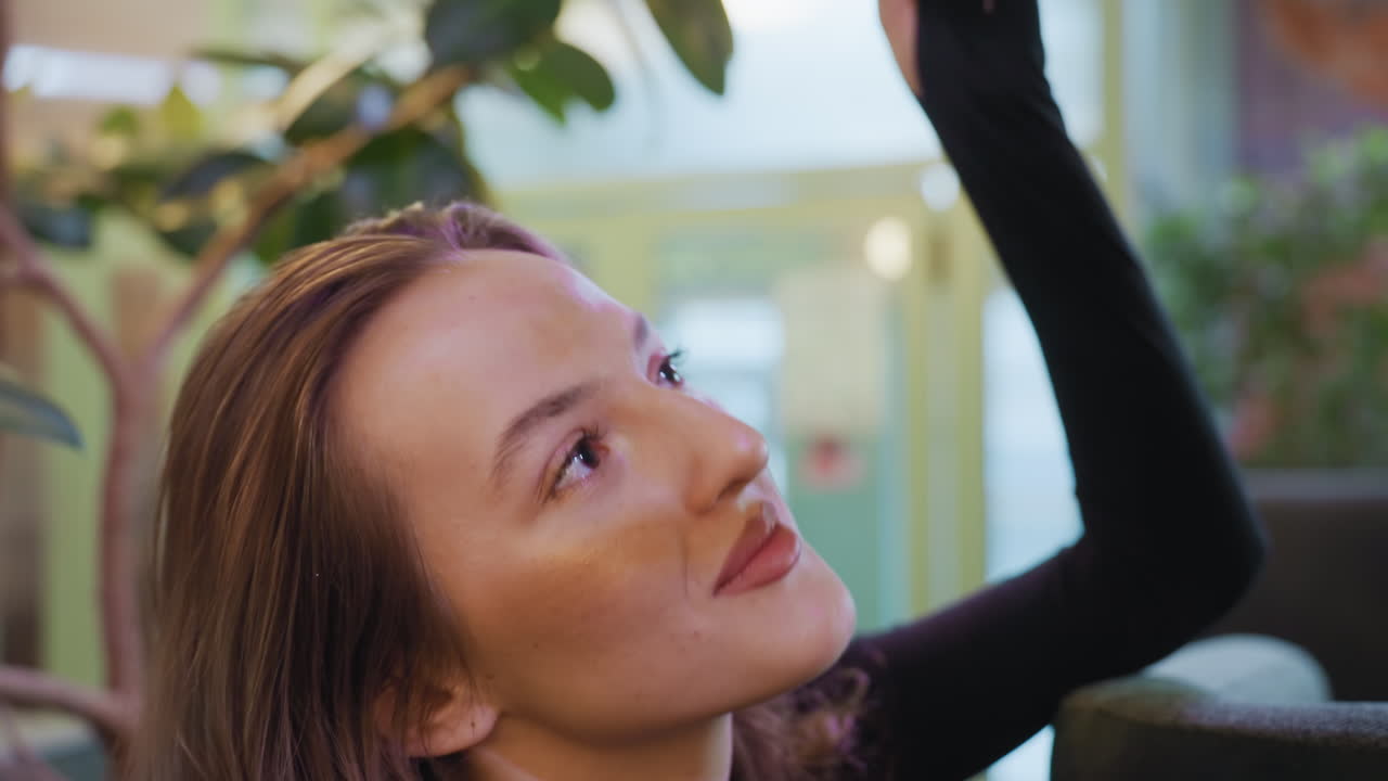 Side profile of young woman with long brown hair reaching up to touch green leaf while resting her arm on cushion, surrounded by lush indoor plants and softly blurred background environment