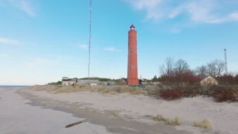 luftaufnahme des roten akmenrags-leuchtturms, ostseeküste, lettland, weißer sandstrand, ruhige see, sonniger tag mit wolken, weit entfernter, aufsteigender drohnenschuss, nach unten geneigt