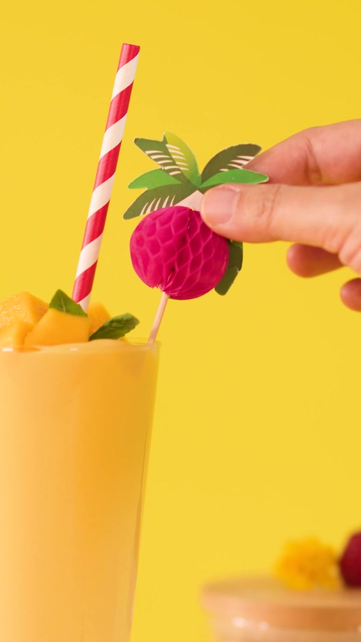 Una mano aggiunge una guarnizione decorativa di frutta di carta rosa a un frullato di mango con cannuccia.