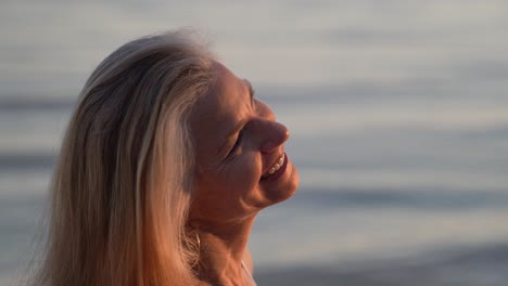 hermosa mujer rubia madura con cabello gris en una playa sonriendo y mirando al sol poniente