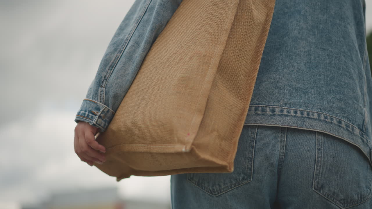 Weiße Frau geht mit einer gewebten Tragetasche in Jeansjacke und Jeans unter bewölktem Himmel, gemütlicher Marktbummel, Nahaufnahme einer Hand, die eine Strohtasche hält, strukturierte Webart und erdige Töne, nachhaltig wiederverwendbar.