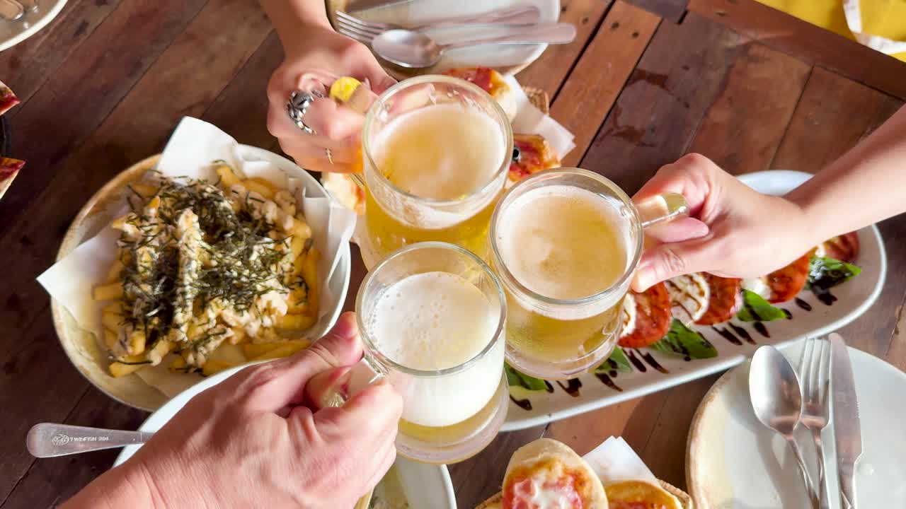 Friends toast with beer over a table of appetizers at a beach club in Phuket, Thailand. Bright, inviting atmosphere