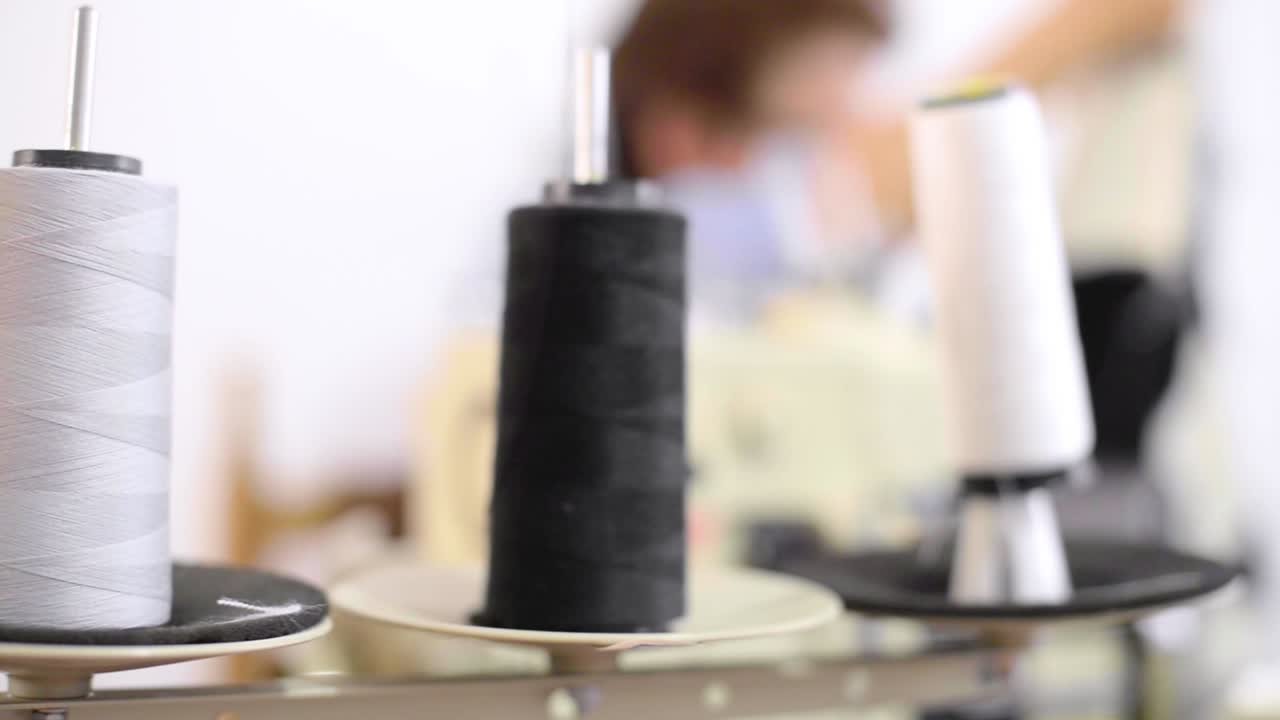 Panning shot of thread's spools on a sewing machine with a latin seamstress working in the background