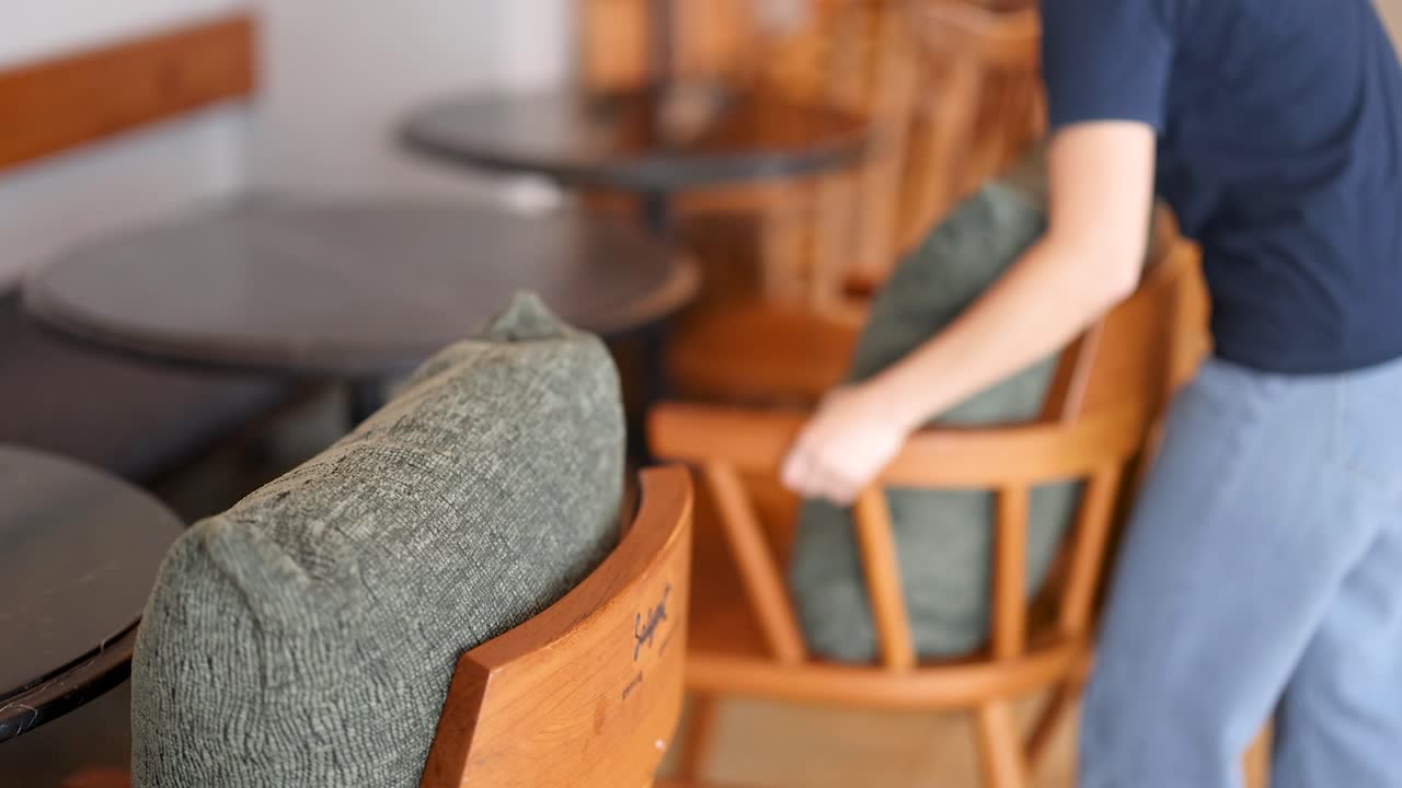 Person arranges green cushion on wooden chair in softly lit, modern cafe with wooden furniture