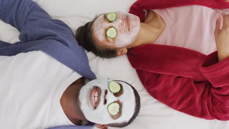 video de una feliz pareja diversa usando máscaras de limpieza y pepino en los ojos acostados en la cama sonriendo