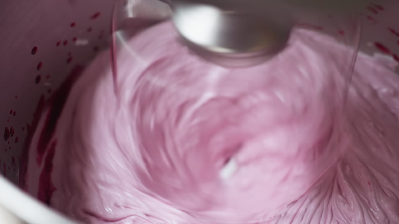 Close up of electric mixer blending flour with red berry puree into smooth pink mixture inside stainless steel bowl, showing swirling motion and creamy texture during vibrant dessert preparation
