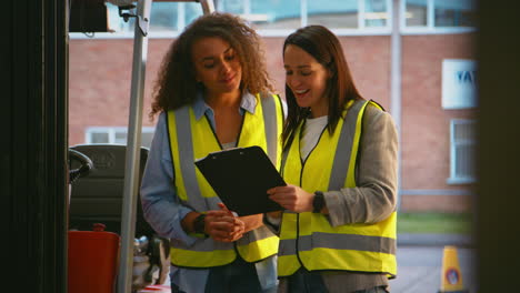 female manager with clipboard in warehouse with female worker standing by fork lift truck