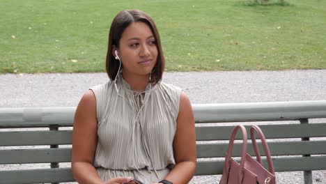 elegant young woman listens to music with her in-ear headphones while sitting on a park bench surrounded by green meadows