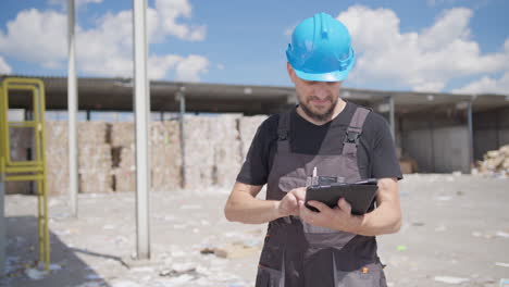 gerente con sombrero azul hace cálculos en una tableta en una instalación de reciclaje de papel