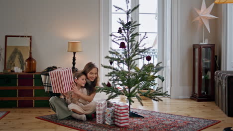 Linda-Familia-Sacudiendo-La-Caja-De-Regalo-Cerca-Del-árbol-De-Navidad.-Mujer-Abrazando-A-Una-Chica-En-Una-Casa-Acogedora