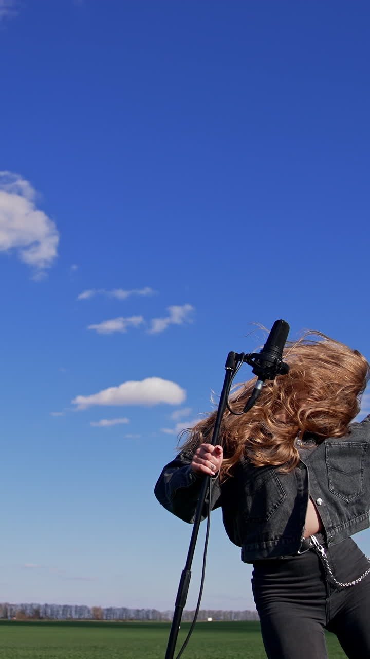 Young rock star dancing near microphone outdoors. Beautiful long-haired girl singing and moving her head with fluttering hair on blue sky background. Vertical video