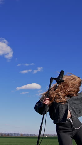 young rock star dancing near microphone outdoors. beautiful long-haired girl singing and moving her head with fluttering hair on blue sky background. vertical video